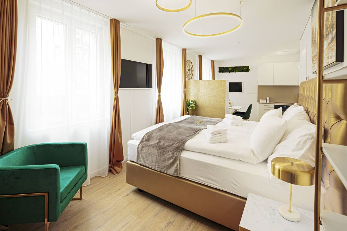 A modern hotel room with a large bed, a green armchair, a small desk, and a kitchen area in the background.