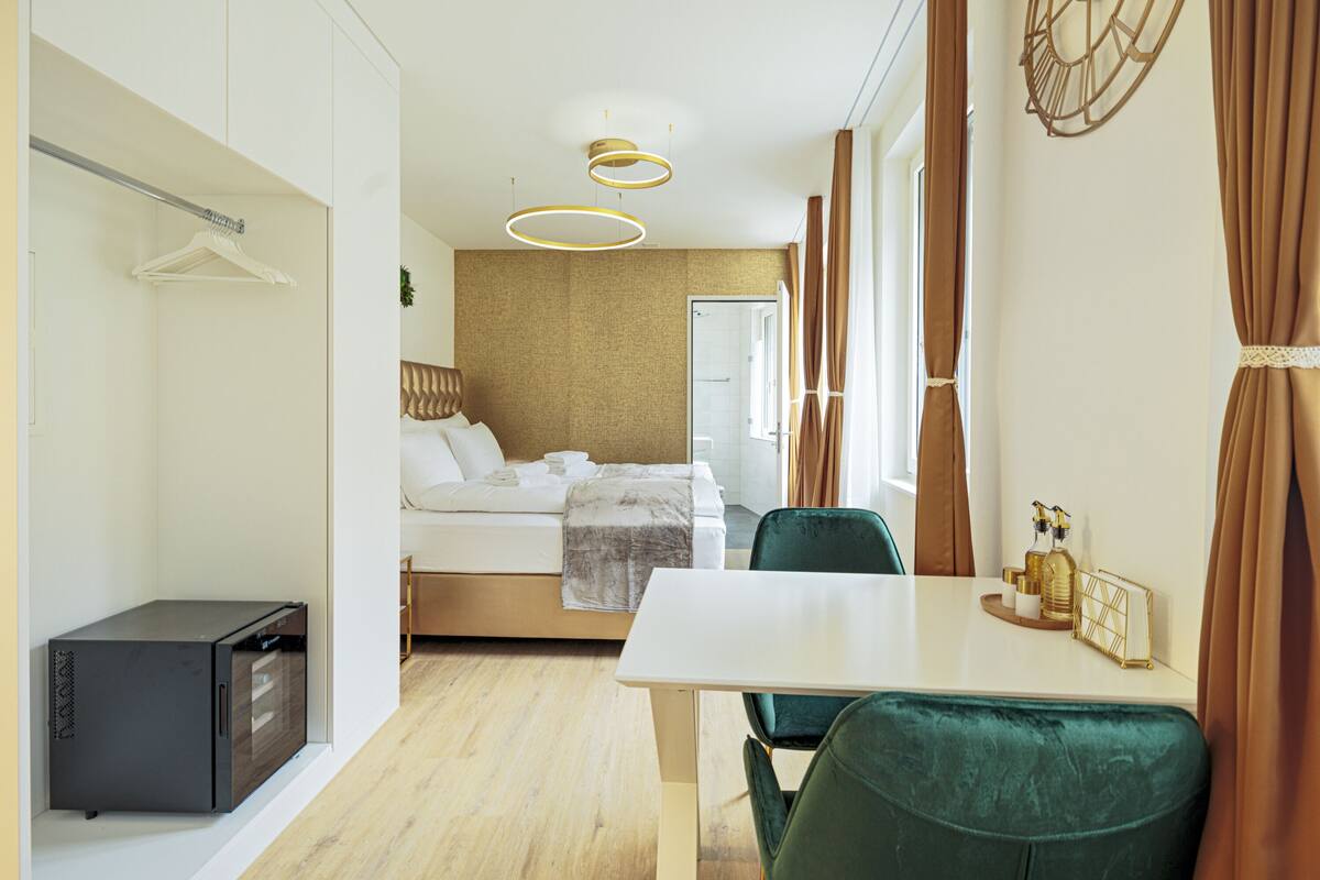 A modern hotel room with a bed, a desk, a chair, a small cabinet, and a shower area.