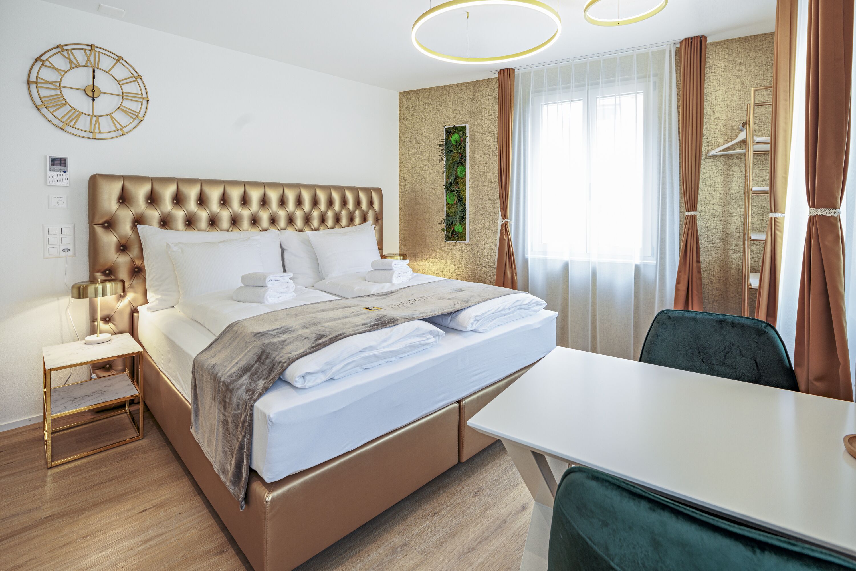Una habitación de hotel moderna con un cabecero capitonado, una cama bien hecha, una mesita de noche con lámpara, un amplio ventanal con cortinas y un armario con una escalera.
