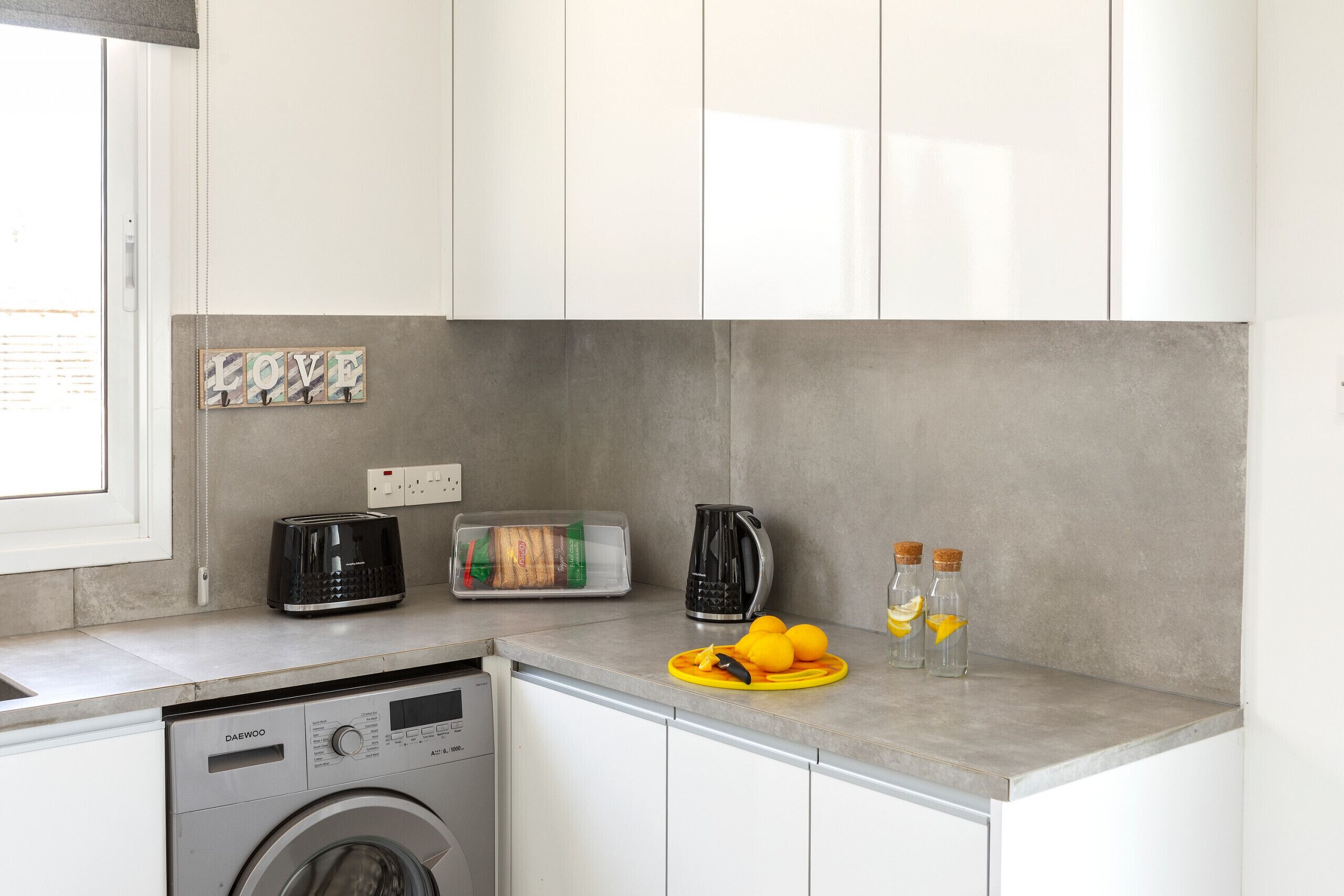Une cuisine équipée d’un lave-linge, d’un grille-pain, d’une bouilloire et d’un plateau contenant du pain et des oranges.
