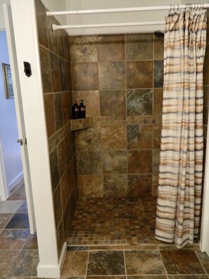 A shower area with stone tiles, a shower curtain, and a shelf with toiletries.