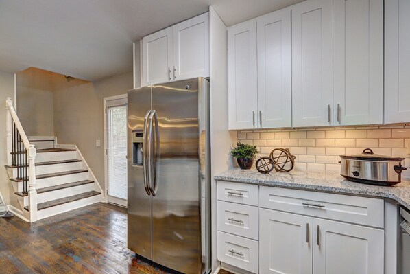 A kitchen with white cabinets, a stainless steel refrigerator, a granite countertop, and a wooden staircase.