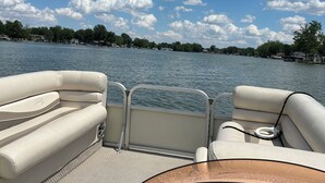 A boat's interior with seating and a control panel, overlooking a calm lake with trees and houses in the distance.