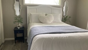 A bedroom with a white bed, a window above the bed, and two bedside tables with plants.