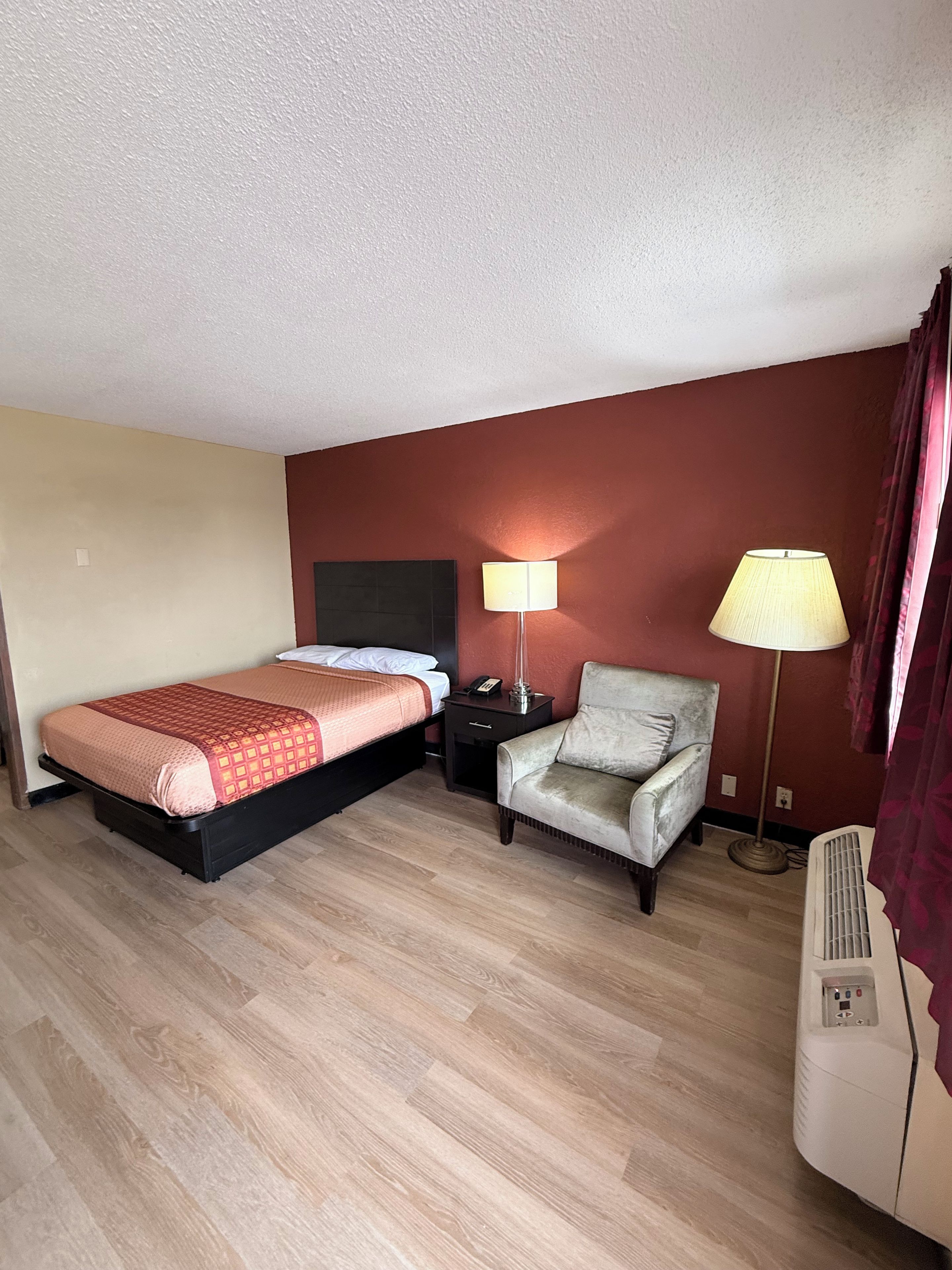 A hotel room with a bed, a chair, a nightstand, a lamp, and an air conditioning unit.