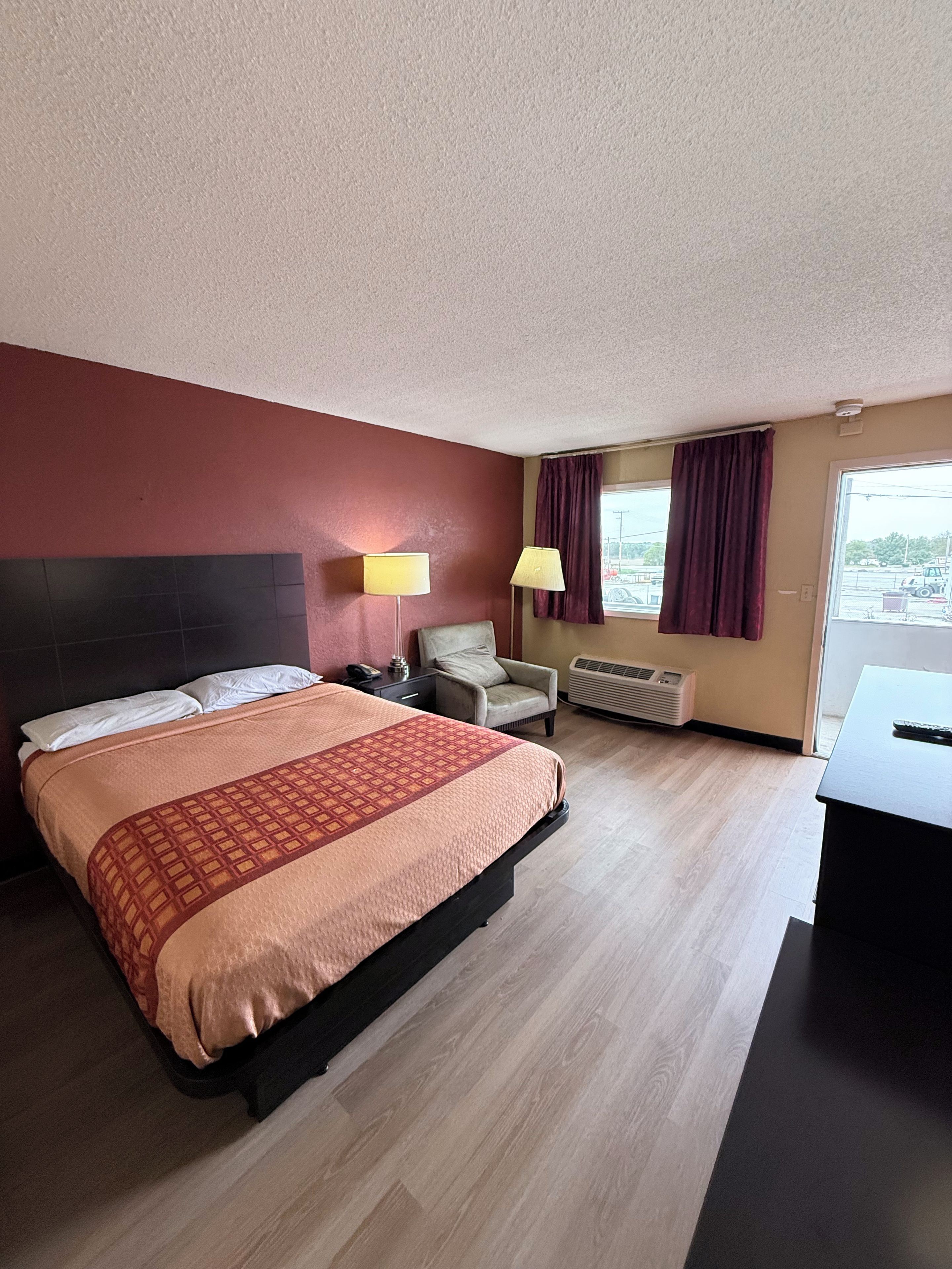 Une chambre d’hôtel avec un lit, un bureau, une chaise, une lampe et une fenêtre avec des rideaux.