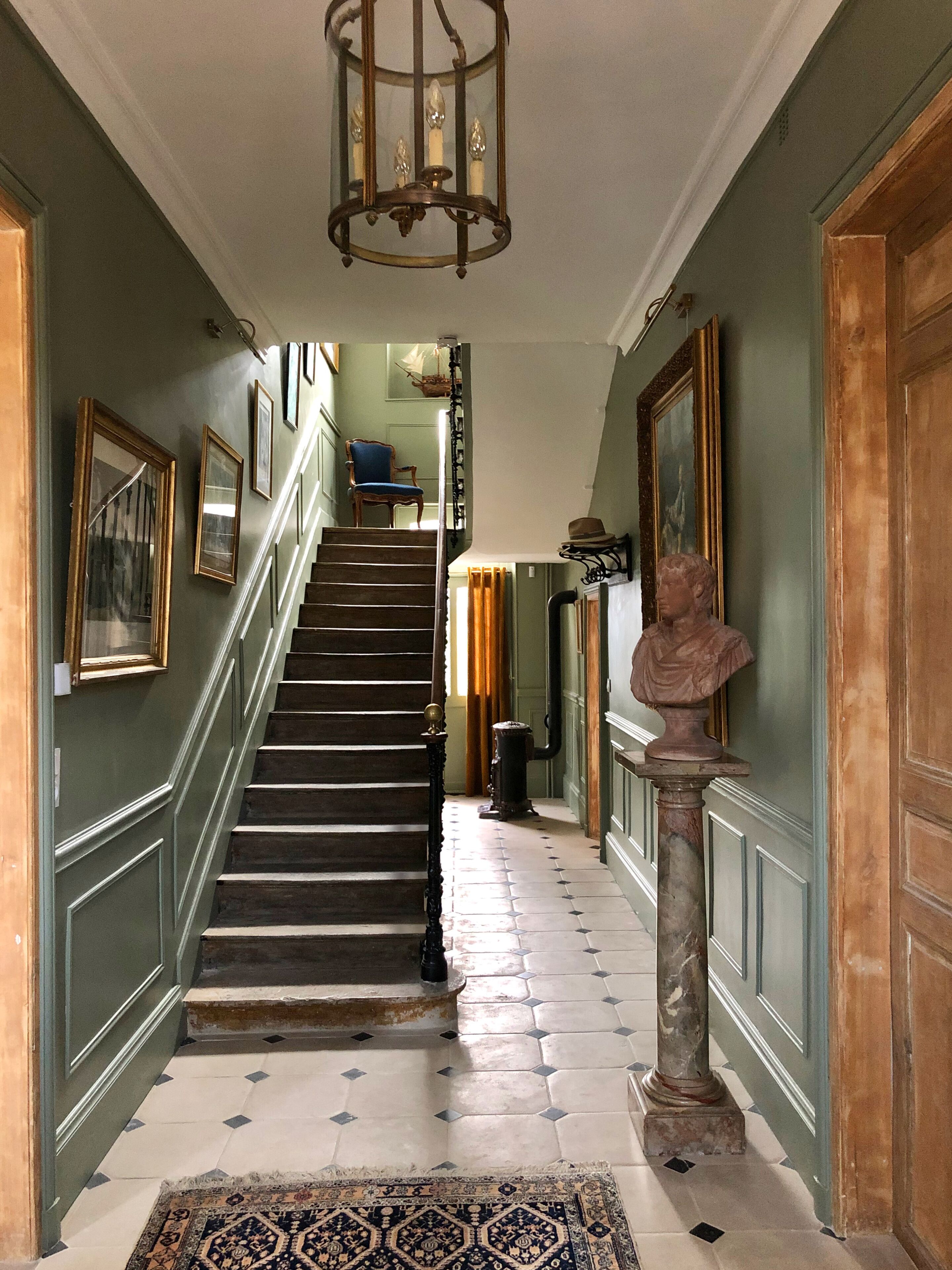 Un grand escalier orné d’une rampe, menant à un palier où se trouvent un buste sculpté et des œuvres d’art encadrées.