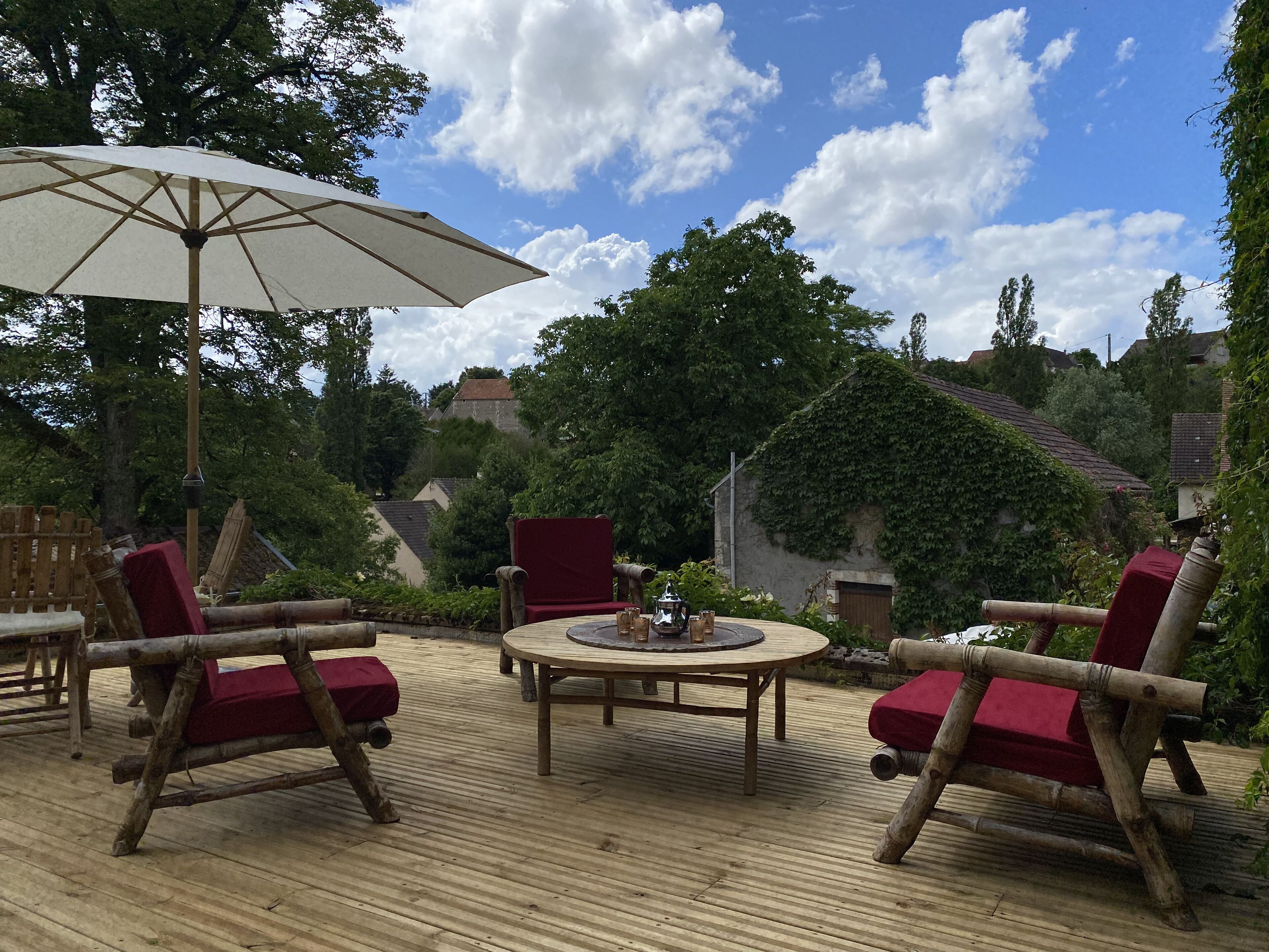 A wooden deck with a round table and red cushioned chairs, an umbrella, and a view of a house and trees.