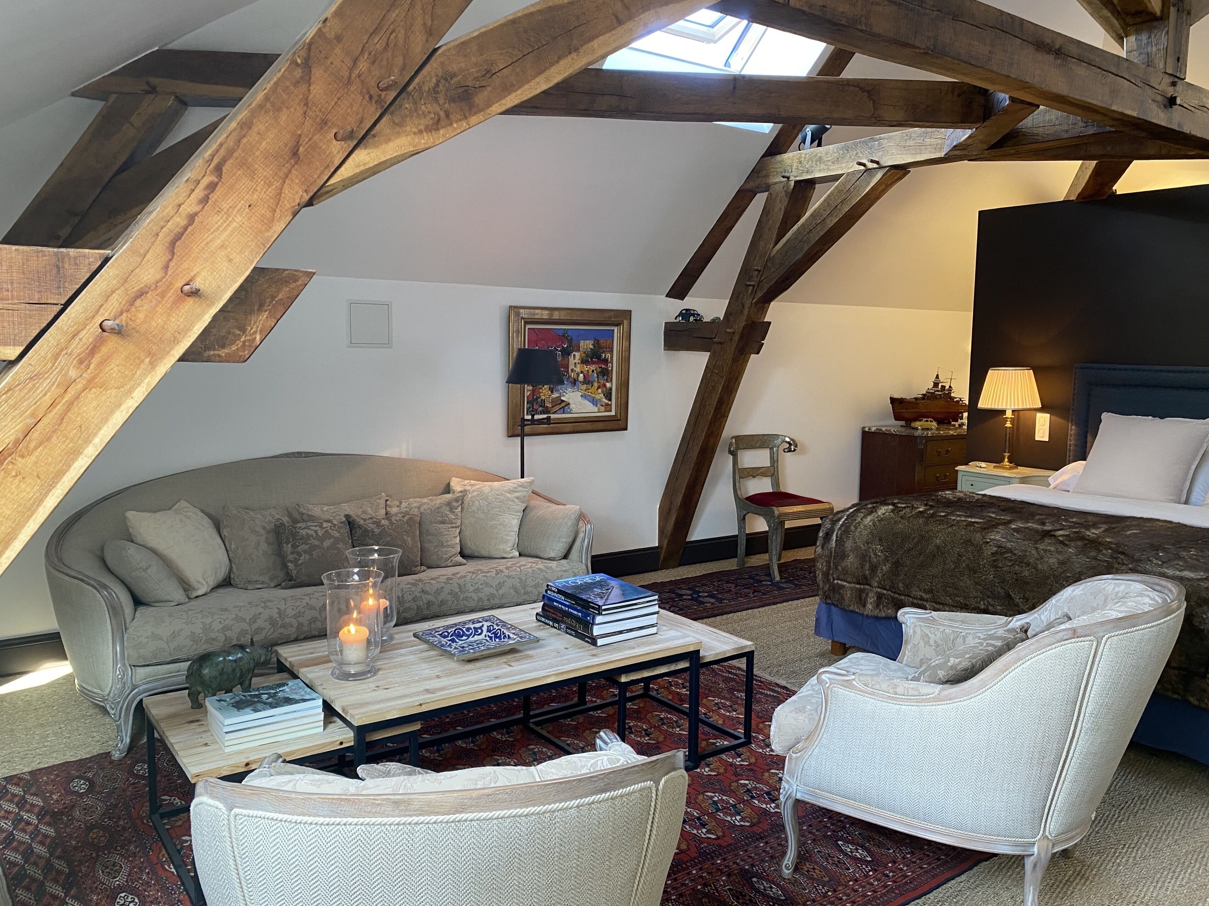 A cozy loft bedroom with exposed wooden beams, a bed, a sofa, a chair, a small table with books and a candle, and a nightstand with a lamp.