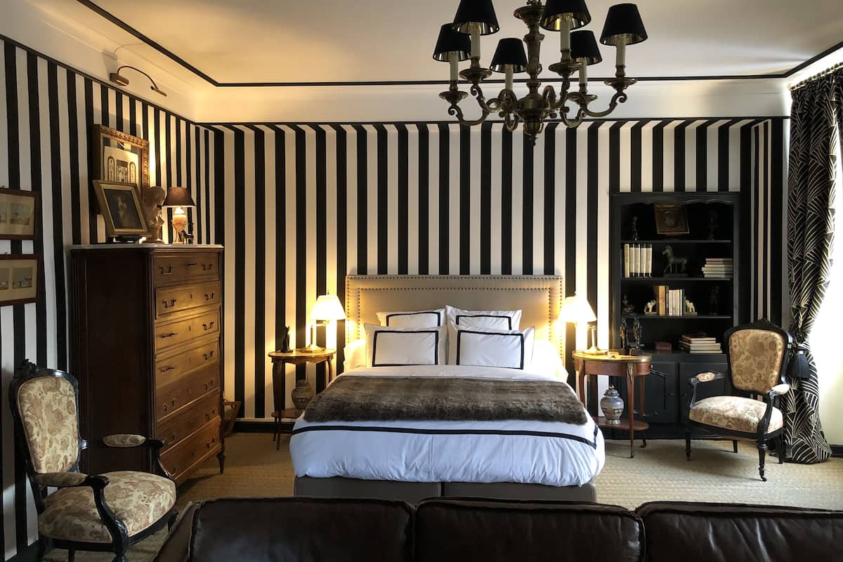 A hotel room with a large bed, a chandelier, a dresser, a bookshelf, and a patterned wall.
