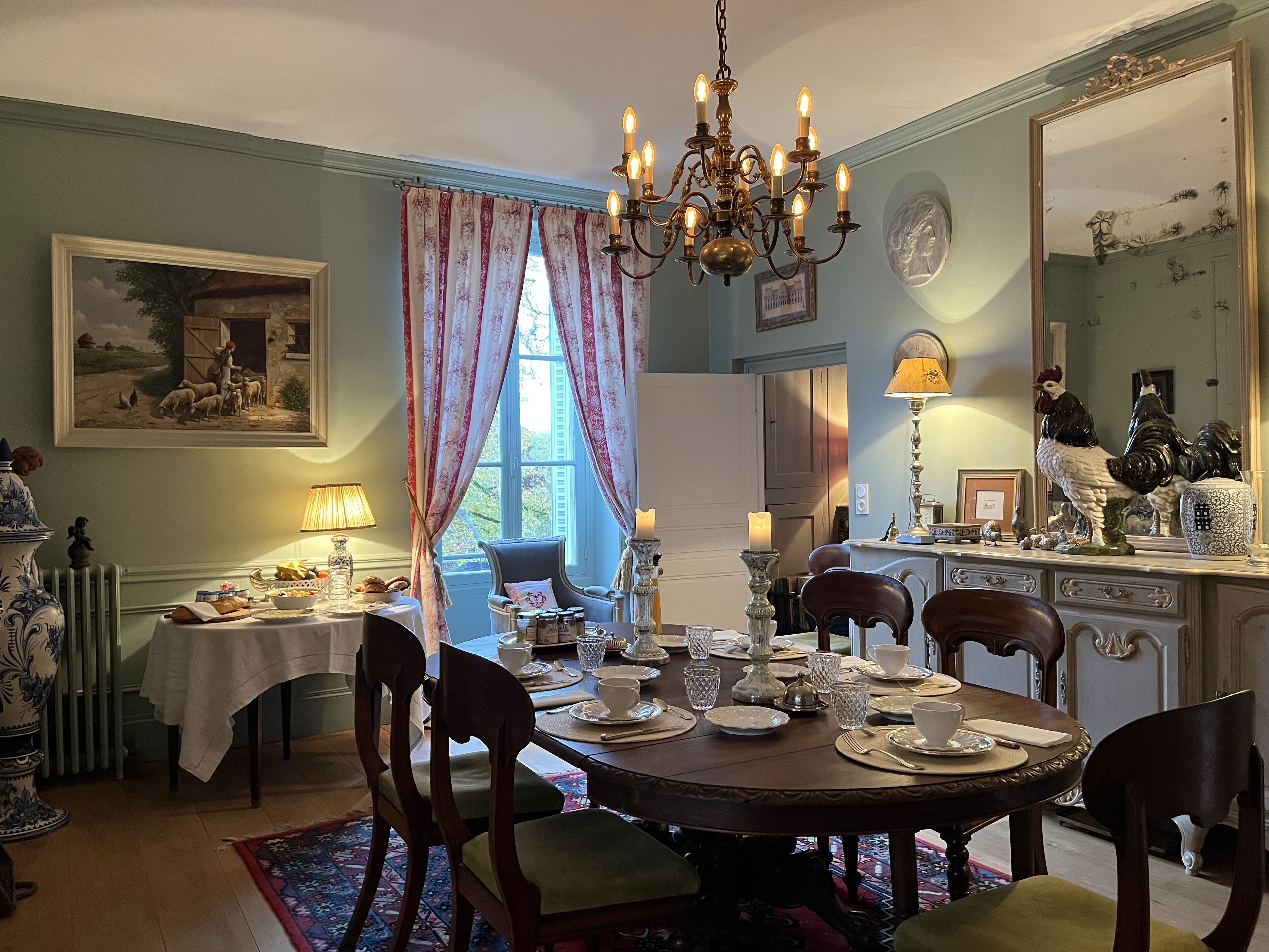 Une salle à manger avec une table ronde dressée pour quatre personnes, entourée de chaises en bois. Un lustre est suspendu au plafond et un grand miroir orne le mur.