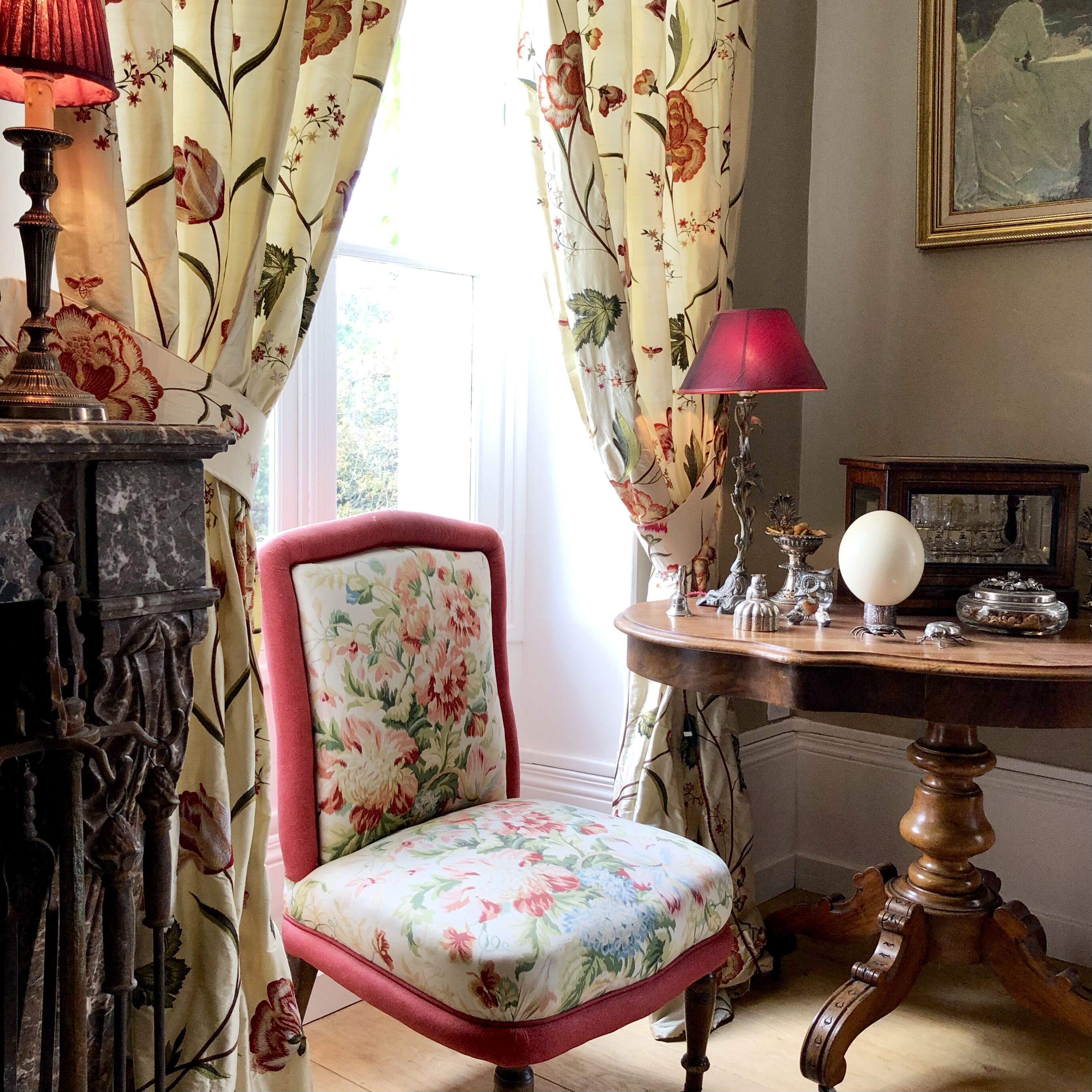 Una habitación con una silla estampada con motivos florales, una mesa con lámpara y un cuadro en la pared.