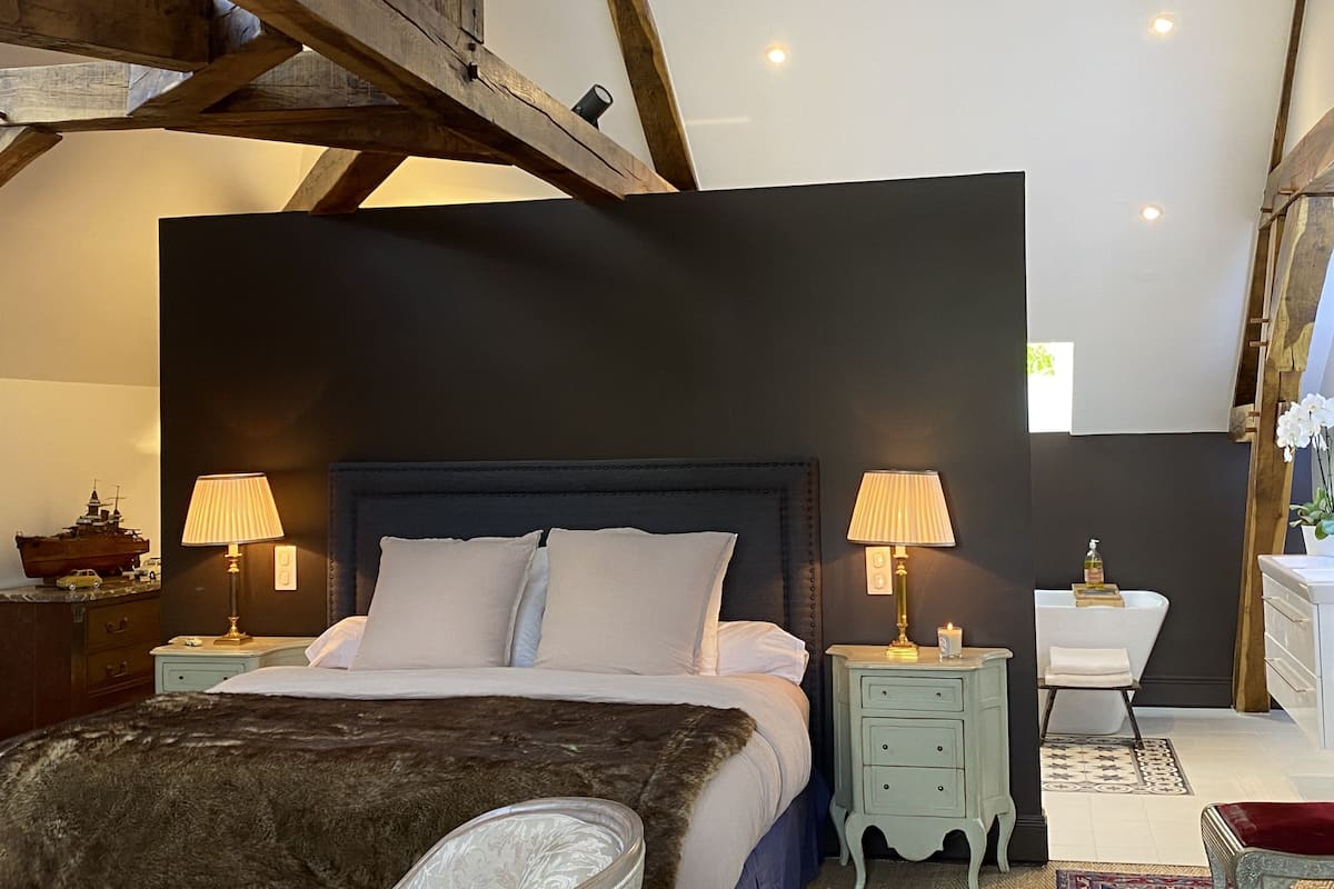 A bedroom with a large bed, two bedside tables with lamps, a nightstand with a ship model, and a small bathroom with a sink and mirror.