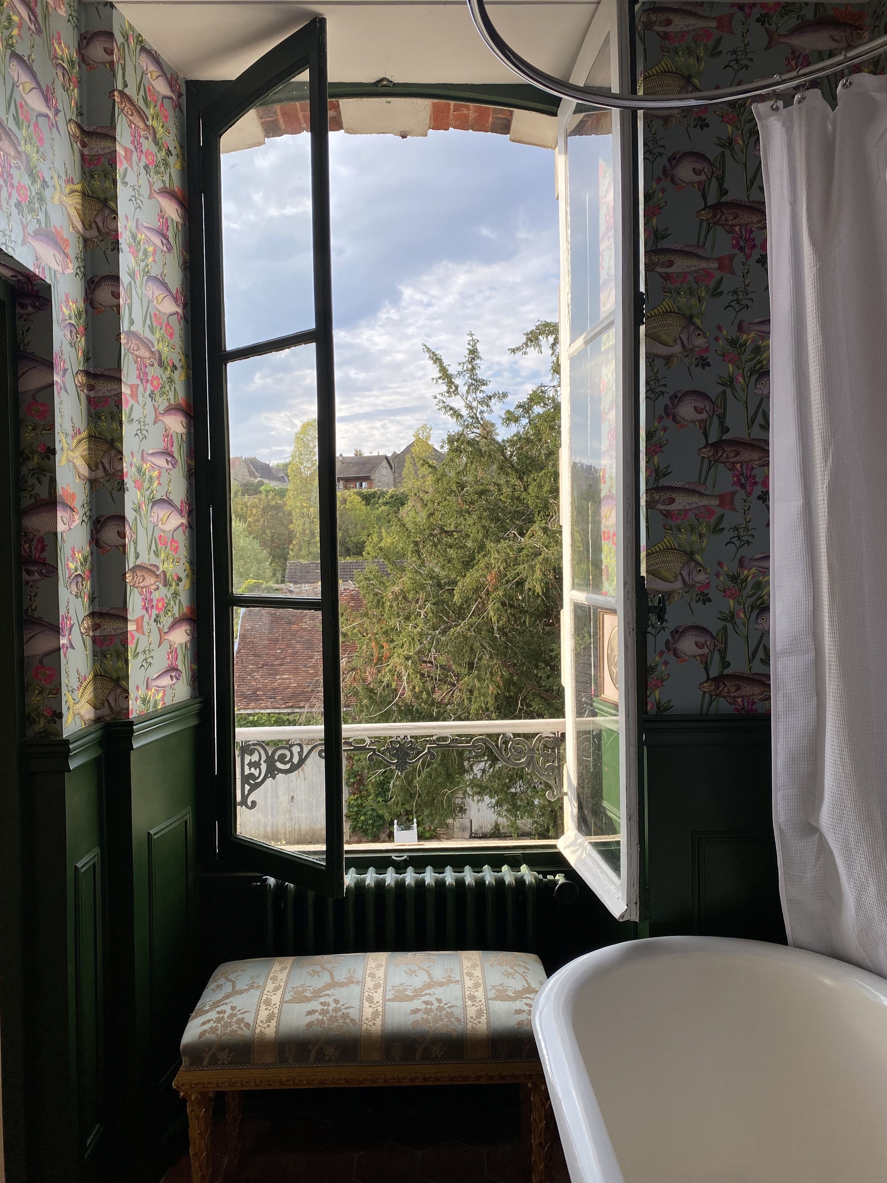 A bathroom with a bathtub, a window with a view of greenery and houses, and patterned curtains.