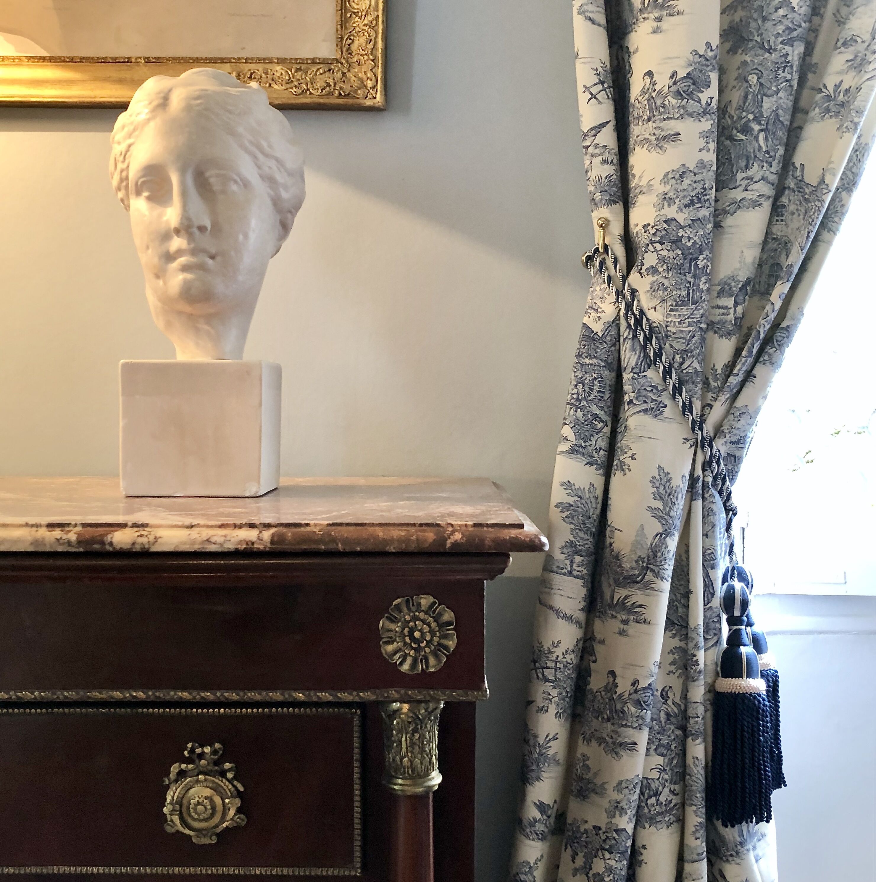 Une sculpture en buste posée sur une commode en bois ornée de motifs complexes, un tableau encadré accroché au mur et des rideaux à motifs floraux.
