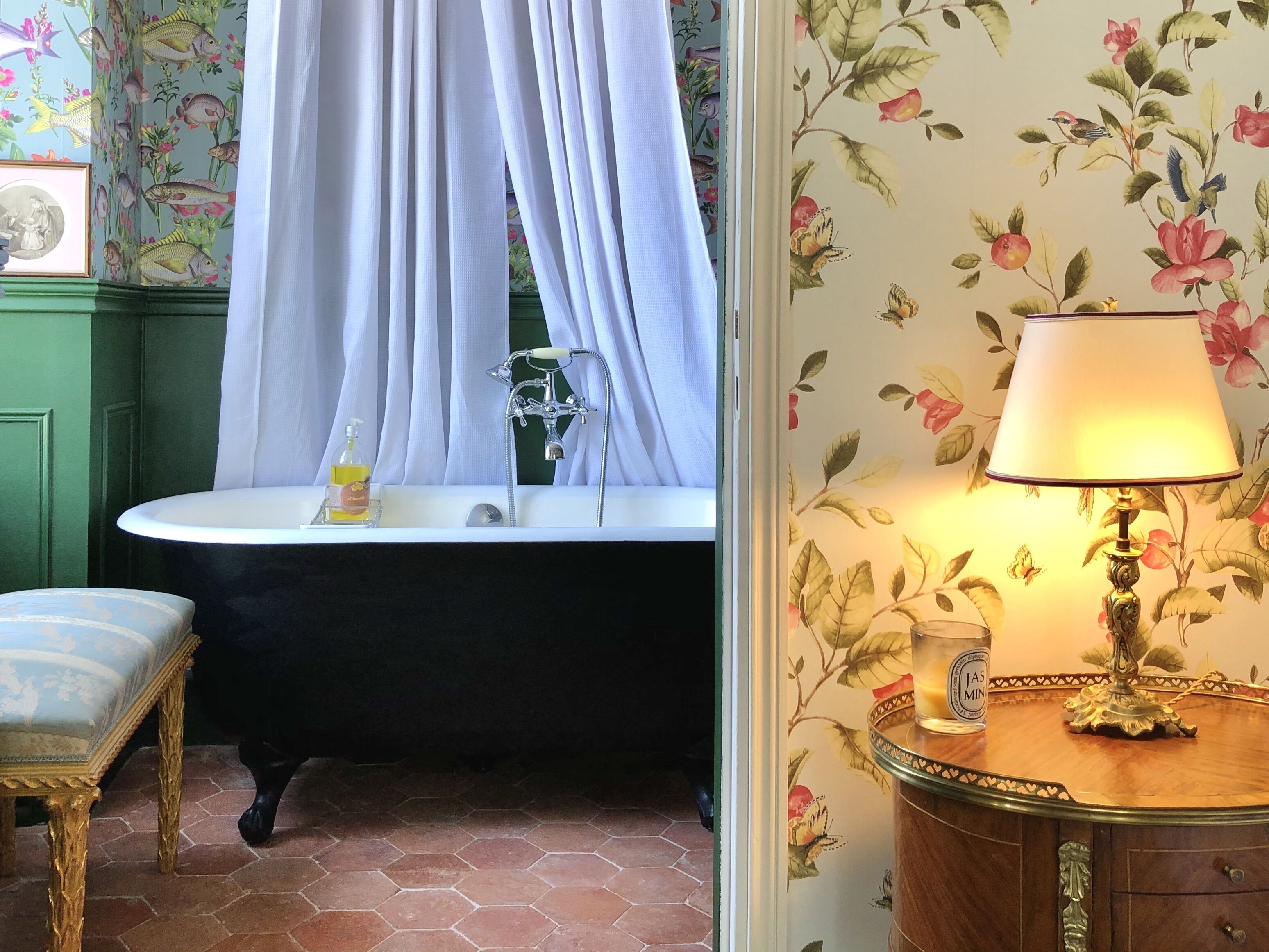 Baño con bañera, lavamanos y espejo. Hay una silla y una mesa con lámpara. Las paredes tienen papel tapiz con motivos florales.