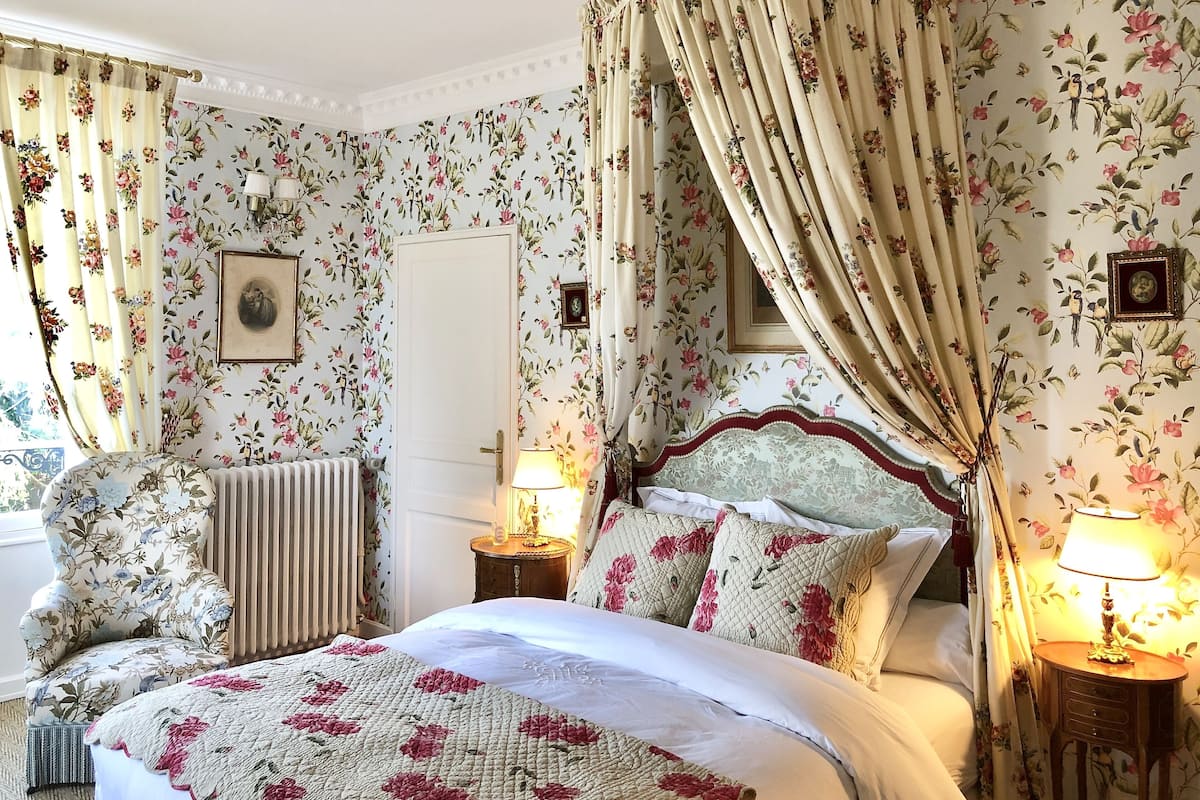 A bedroom with floral wallpaper, a canopy bed, two bedside tables with lamps, a radiator, and a chair.