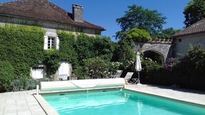 Une piscine dotée d’une terrasse en pierre, entourée d’une végétation luxuriante et bordée d’un bâtiment en pierre surmonté d’un toit en tuiles.