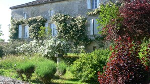 Un bâtiment en pierre, doté de volets blancs, entouré d’une végétation luxuriante et de plantes en fleurs.