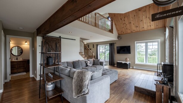 A spacious living area with a large gray sectional sofa, wooden flooring, a wooden staircase, a TV mounted on the wall, and a fireplace.