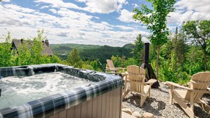 A hot tub surrounded by Adirondack chairs, a chimney, and a scenic view of mountains and trees.