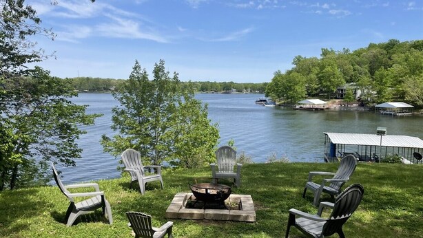 A lakeside outdoor seating area with Adirondack chairs and a fire pit, surrounded by lush greenery and a view of the lake. - WATERFRONT Vacation! {3 bed 2 bath.  Sleeps 13} (Goreville)