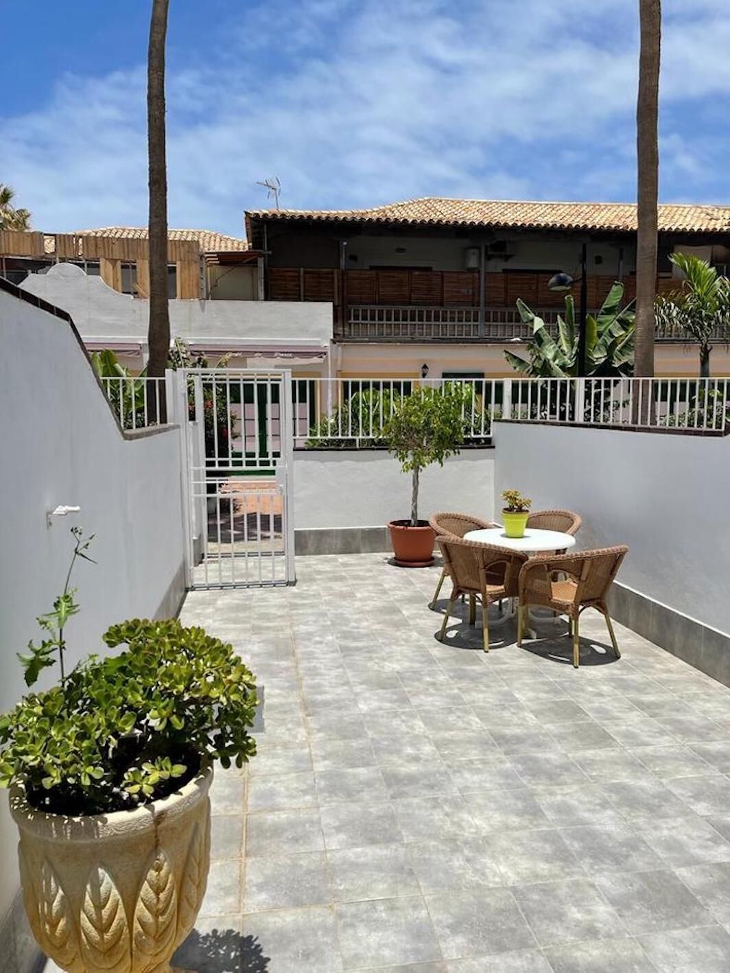 Un patio en la azotea con una mesa y sillas, rodeado de plantas en macetas y una cerca blanca.