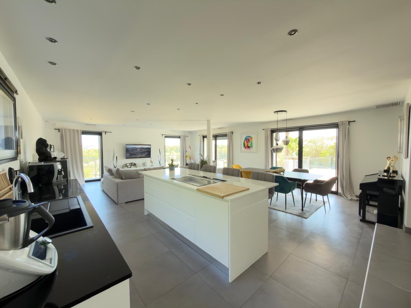 A modern kitchen with a central island, dining area with chairs, and a living space with a sofa and TV.