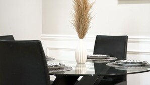 Una mesa de comedor con sillas negras, un florero blanco con plantas secas y platos blancos y negros sobre individuales.