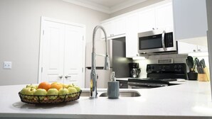 Una cocina moderna con armarios blancos, un lavamanos de acero inoxidable y un frutero con manzanas y naranjas.