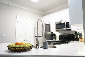 A modern kitchen with white cabinets, a stainless steel sink, and a fruit bowl containing apples and oranges.