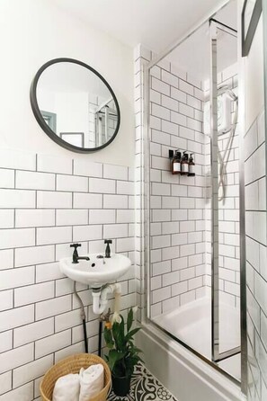 Un baño con paredes de azulejos blancos, un lavamanos blanco, un espejo y una cabina de ducha de vidrio.