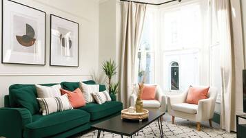 Una sala de estar con un sofá verde, un sillón blanco y una mesa de centro. Hay dos cuadros abstractos enmarcados en la pared y una ventana con cortinas.