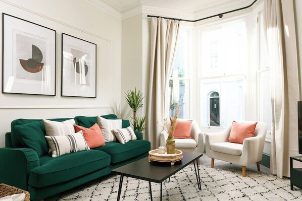 A living room with a green sofa, white armchair, and a coffee table. There are two framed abstract paintings on the wall and a window with curtains.