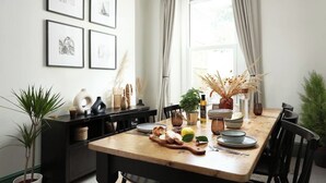 A dining area with a wooden table, chairs, and a window with curtains. There are framed pictures on the wall, a potted plant, and a vase with dried plants on the table. - Luxury for 2024, Central Plymouth Home for 10 (Plymouth)