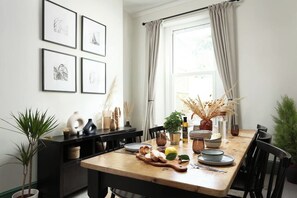 A dining area with a wooden table, chairs, and a window with curtains. There are framed pictures on the wall, a potted plant, and a vase with dried plants on the table.