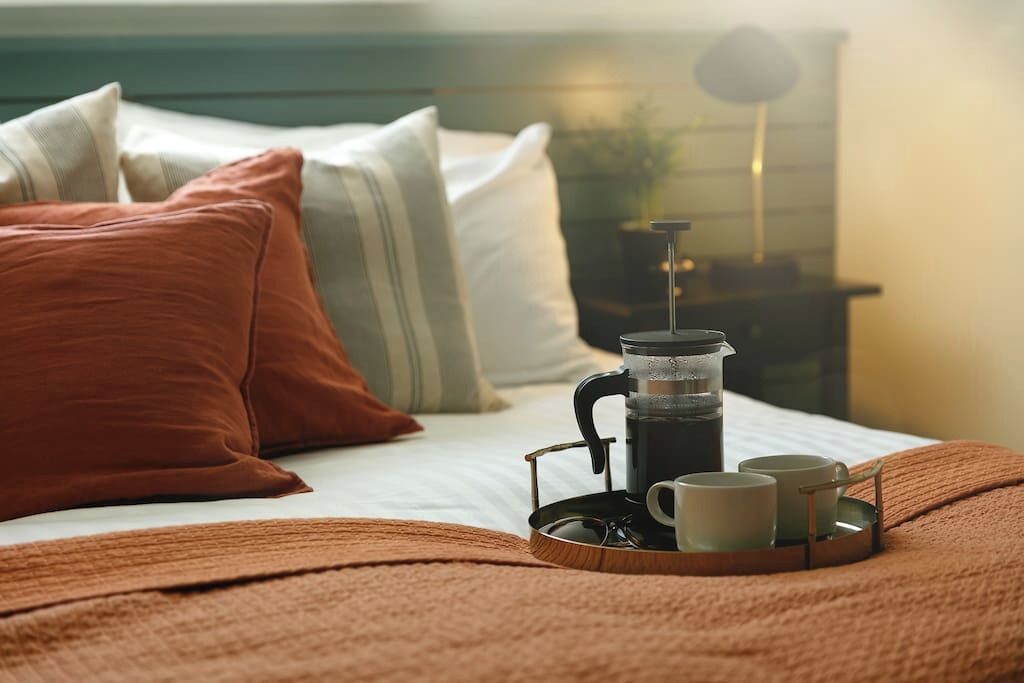 A neatly made bed with a coffee maker, two cups, and a tray on a wooden bedspread.