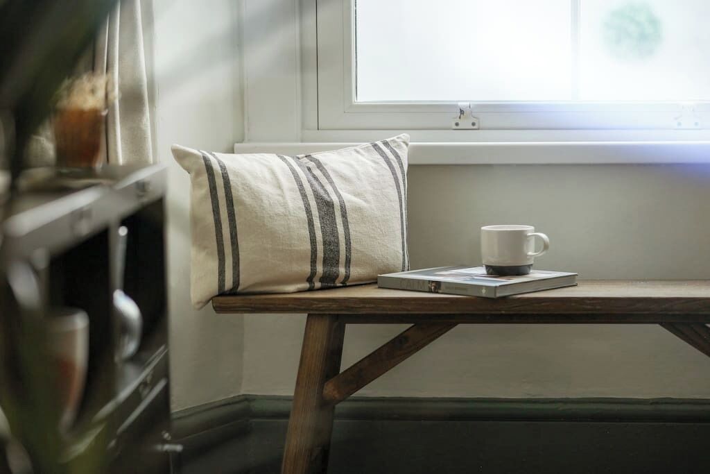A small wooden table with a white mug and a striped pillow, positioned near a window.