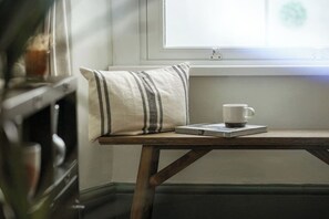 Une petite table en bois, sur laquelle sont posés une tasse blanche et un coussin à rayures, est placée près d’une fenêtre.