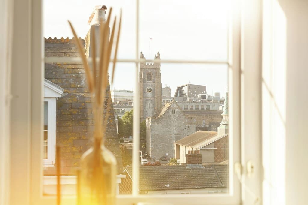 Vista a través de una ventana que muestra un edificio histórico con una torre de reloj, un muro de ladrillo y una estructura moderna al fondo.