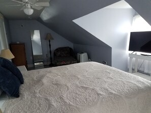 A bedroom with a large bed, a desk with a computer, a ceiling fan, and a window.