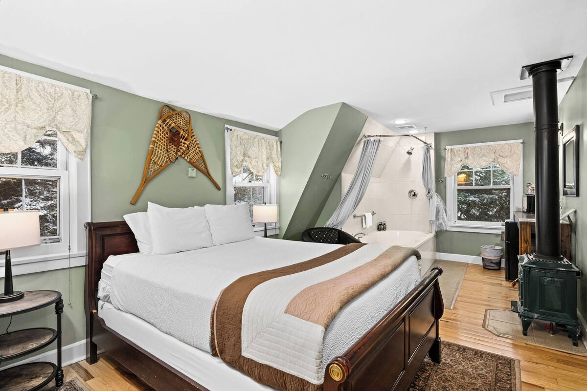 A bedroom with a large bed, a wooden headboard, a nightstand, a lamp, a window with curtains, a shower area, and a wood-burning stove.