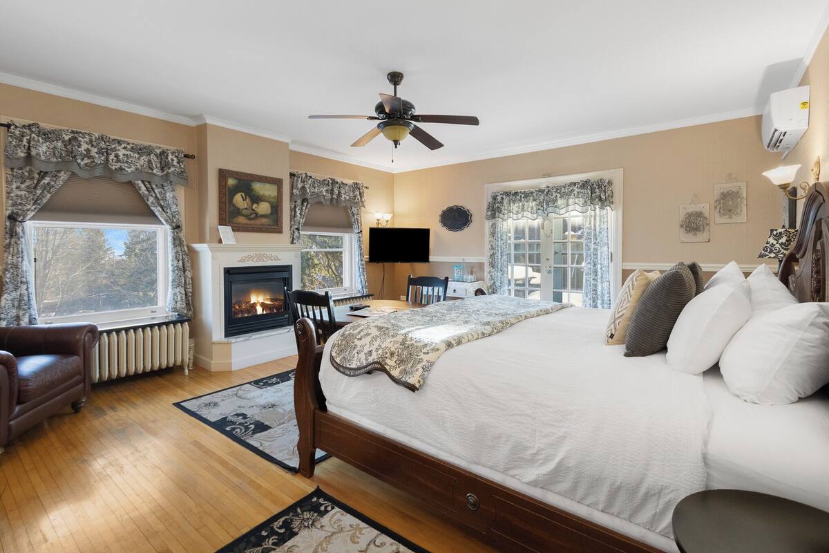 A bedroom with a large bed, a fireplace, a ceiling fan, and a view of the outdoors.