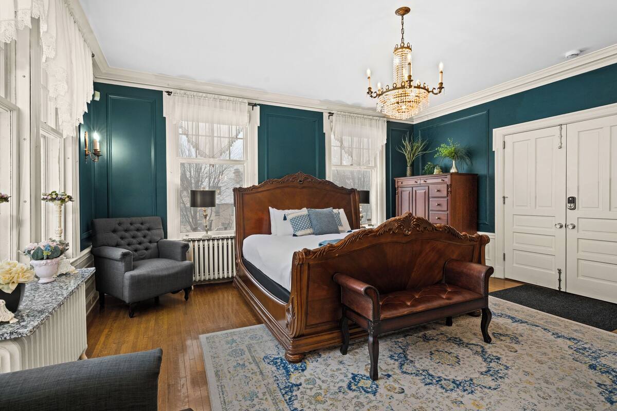 A bedroom with a four-poster bed, a leather armchair, a wooden dresser, and a chandelier.