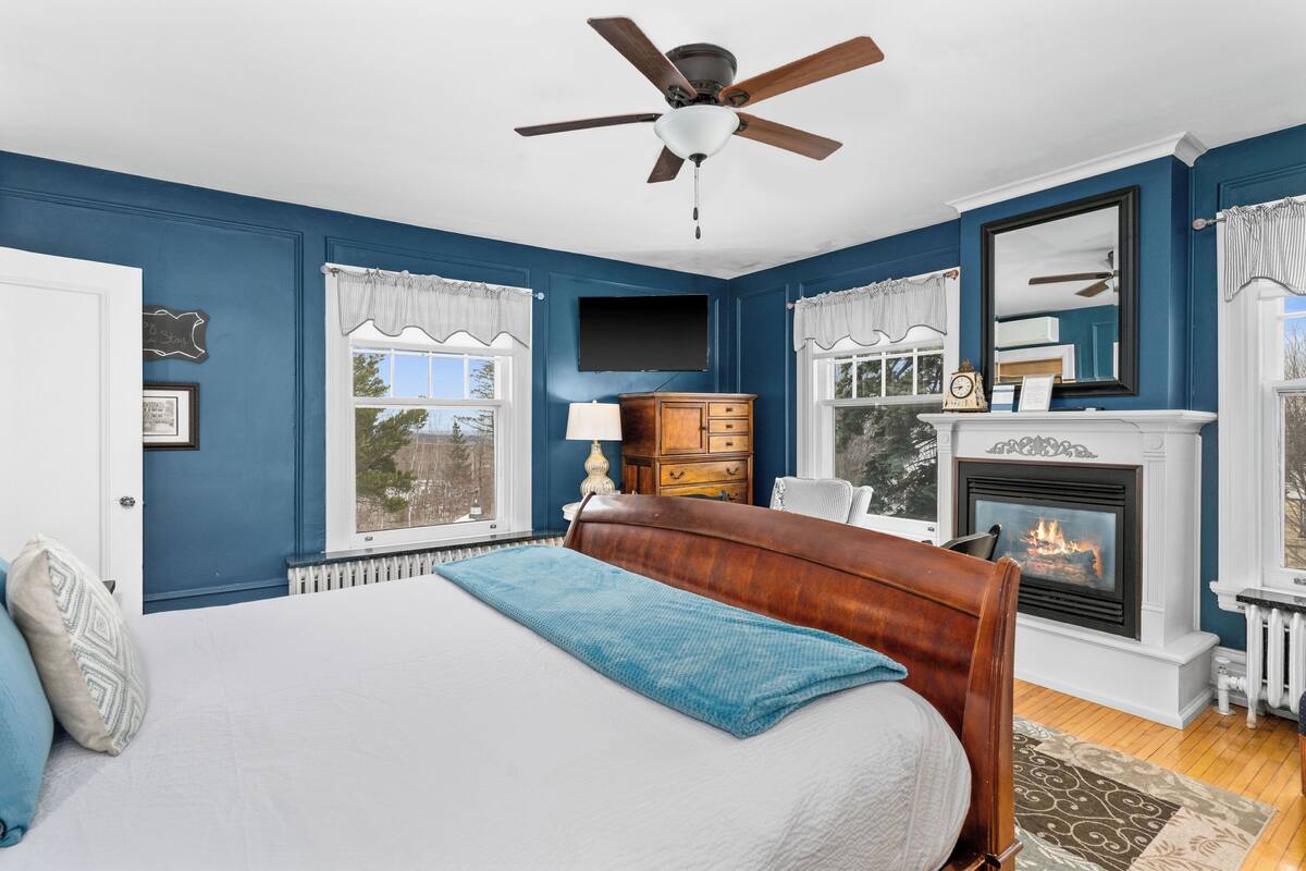 A bedroom with a large bed, a ceiling fan, a fireplace, and a television mounted on the wall.
