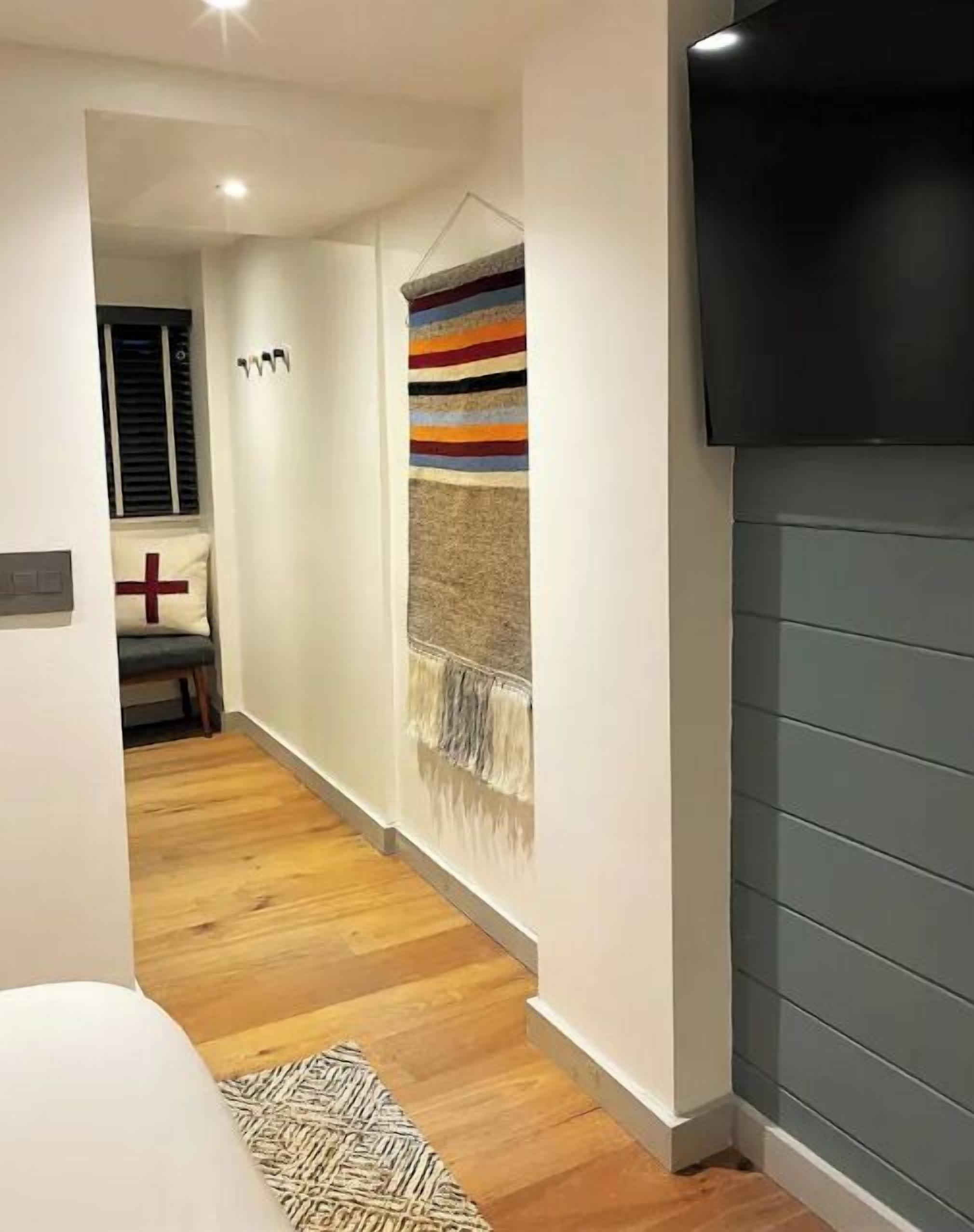 Un estrecho pasillo con suelo de madera, una cama con cabecera blanca, un televisor montado en la pared y un tapiz a rayas.