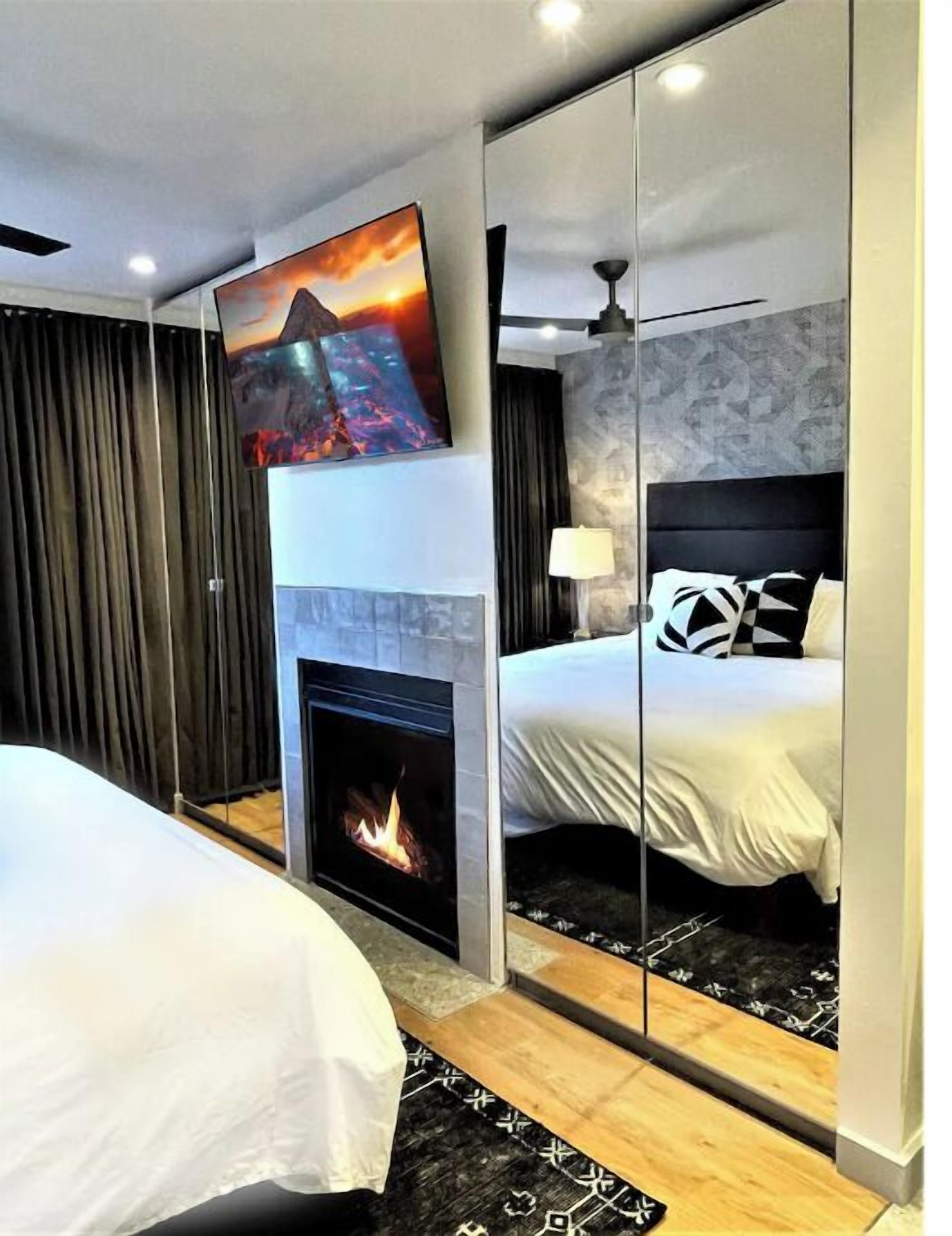 A modern hotel room with a fireplace, a bed with black and white bedding, a TV mounted on the wall, and a large mirror reflecting the room.