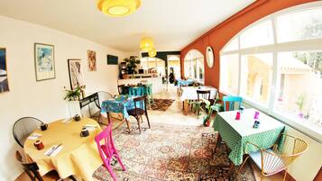 A dining area with multiple tables and chairs, a patterned rug, and large windows with a view of the outside.