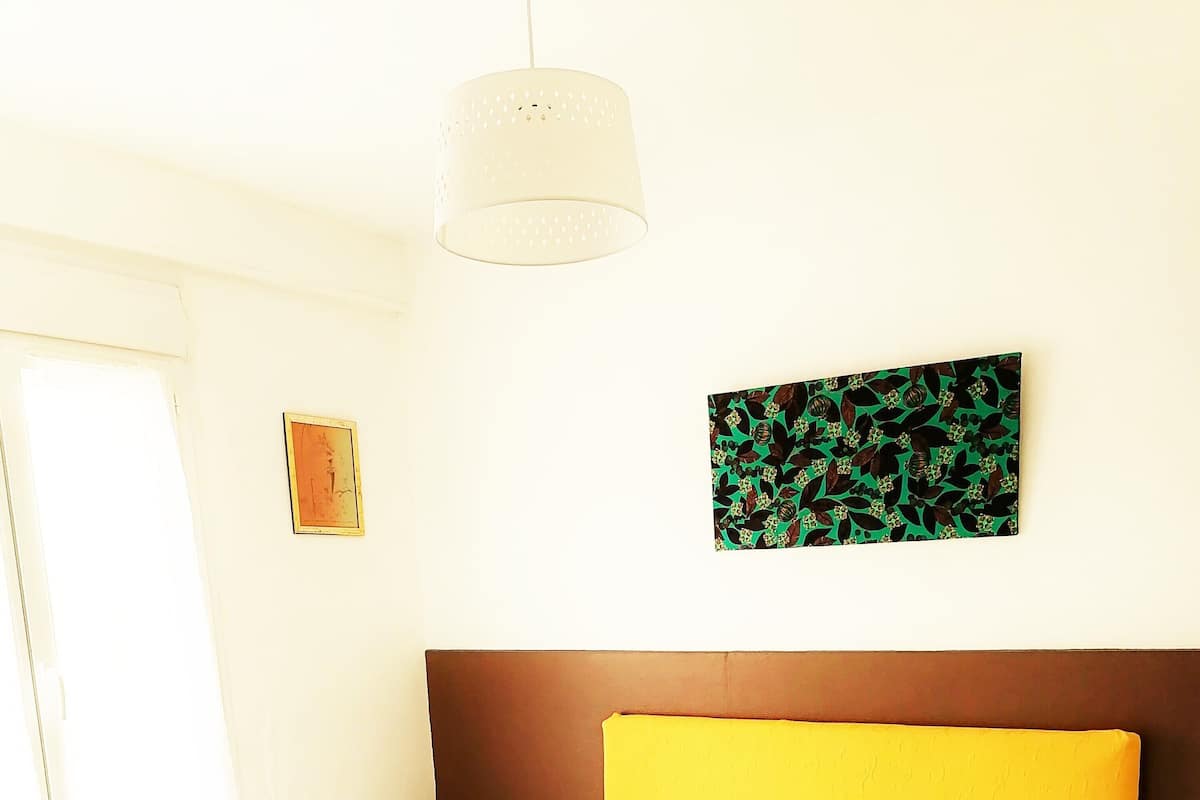Un lit bien fait, avec du linge de lit blanc, une table de chevet en bois surmontée d’une lampe blanche et un tableau accroché au mur.