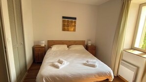Un letto rifatto con asciugamani, un comodino con una lampada, una finestra con tende e un termosifone.
